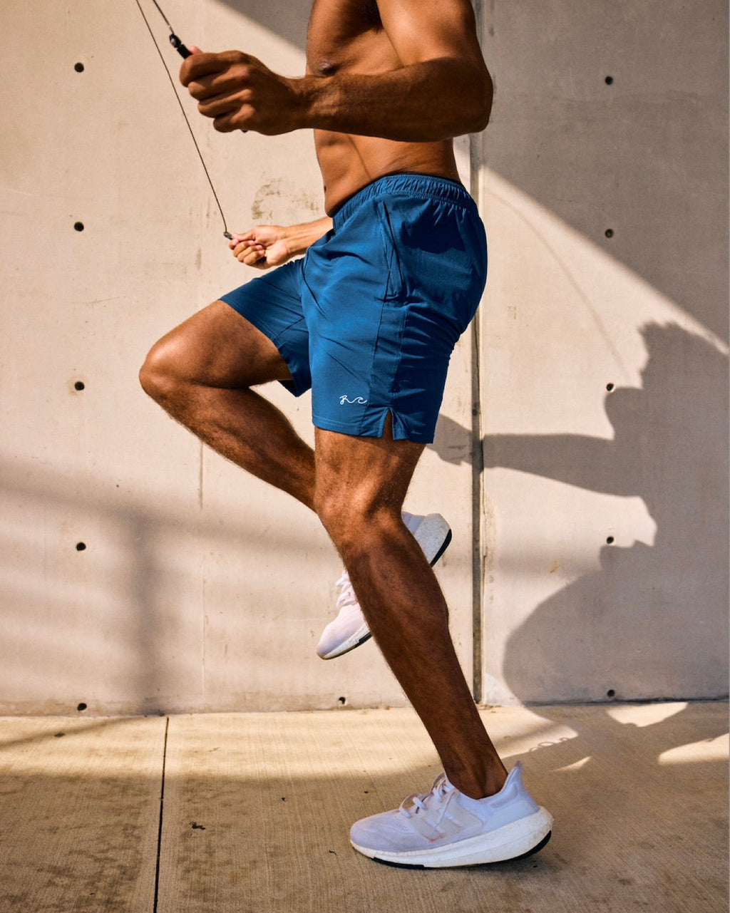 A shirtless man in blue athletic shorts and white sneakers jumps rope against a concrete wall with strong sunlight and shadows.