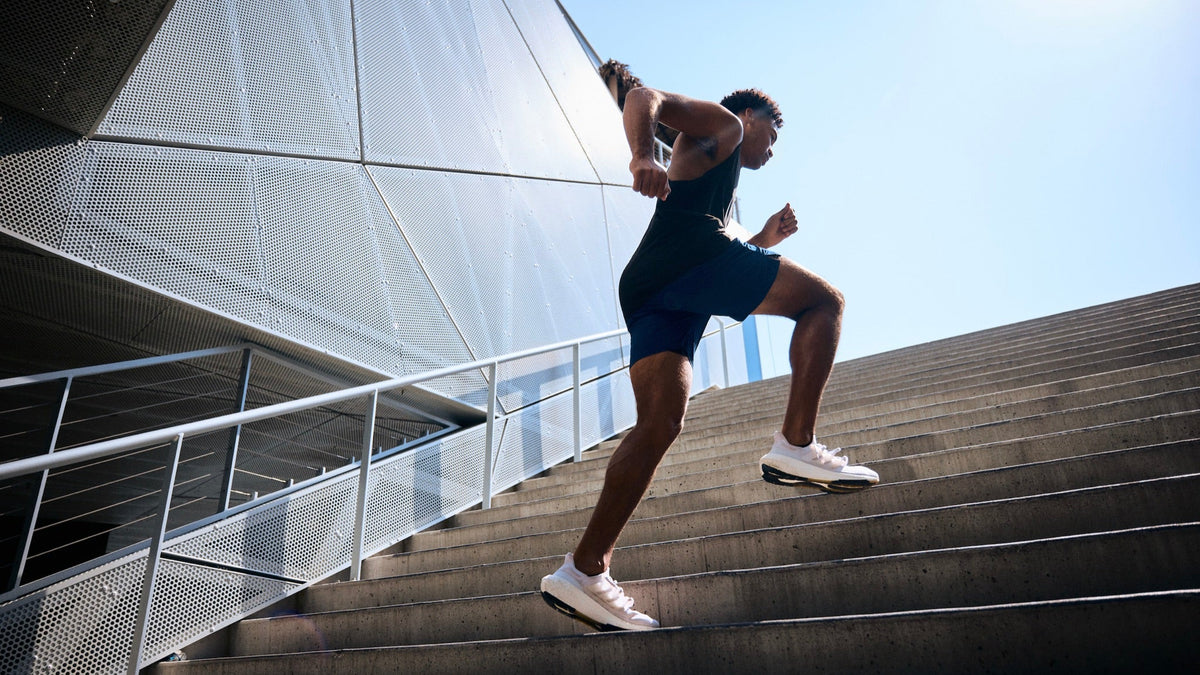 A man in a dark blue athletic outfit and white sneakers running up outdoor concrete stairs under a bright sky.