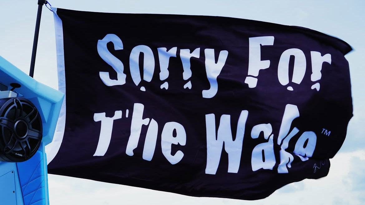 A black flag with the phrase 'Sorry For The Wake' in large white stylized lettering, shown outdoors against a pale sky. Part of a blue structure is visible on the left.