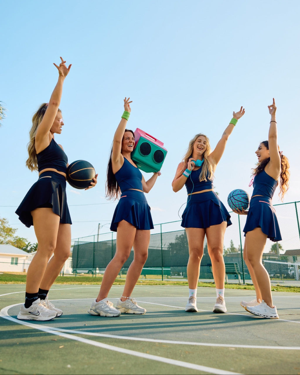 Four women in matching navy blue athletic outfits pose on an outdoor basketball court under a clear blue sky. Two hold basketballs, and one holds a retro green boombox.