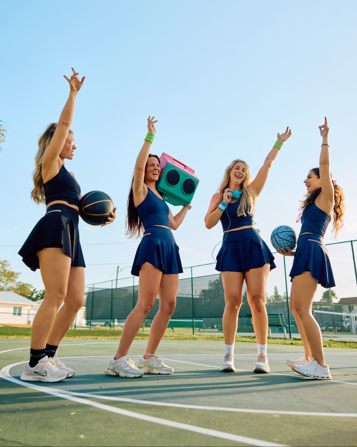 Four women in matching navy blue athletic outfits pose on an outdoor basketball court under a clear blue sky. Two hold basketballs, and one holds a retro green boombox.