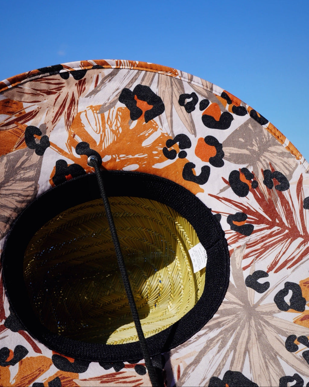 A close-up of a wide-brimmed sun hat with a tropical floral and leopard print pattern in orange, black, and beige, photographed against a clear blue sky.