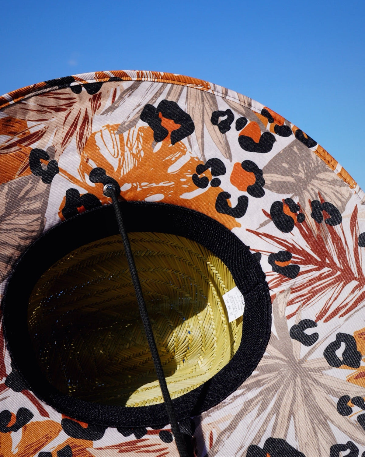 A close-up of a wide-brimmed sun hat with a tropical floral and leopard print pattern in orange, black, and beige, photographed against a clear blue sky.