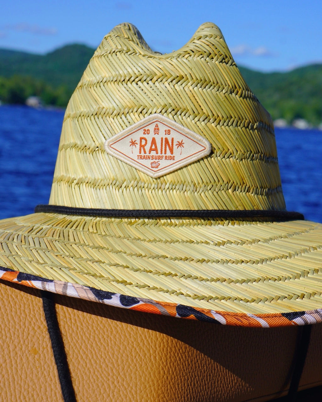 A straw wide-brim hat with a tan leather patch reading 'RAIN TENSFOOT CO.' in orange, photographed outdoors with blue water and green hills in the background.