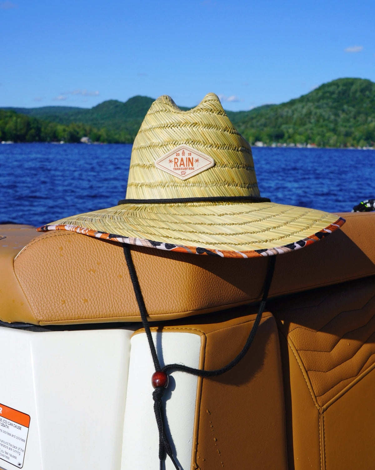 A straw cowboy-style sun hat with a tan woven crown and a wide brim, resting on the back of a tan boat seat. The hat has a visible 'The Rain' logo patch on the front and a patterned underside to the brim, with a blue lake and green hills in the background.