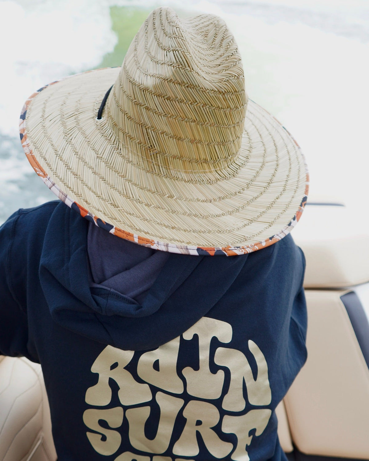 A person wearing a navy blue hoodie with a large beige 'RAIN SURF SHOP' graphic on the back, seated in a vehicle and wearing a straw sun hat.
