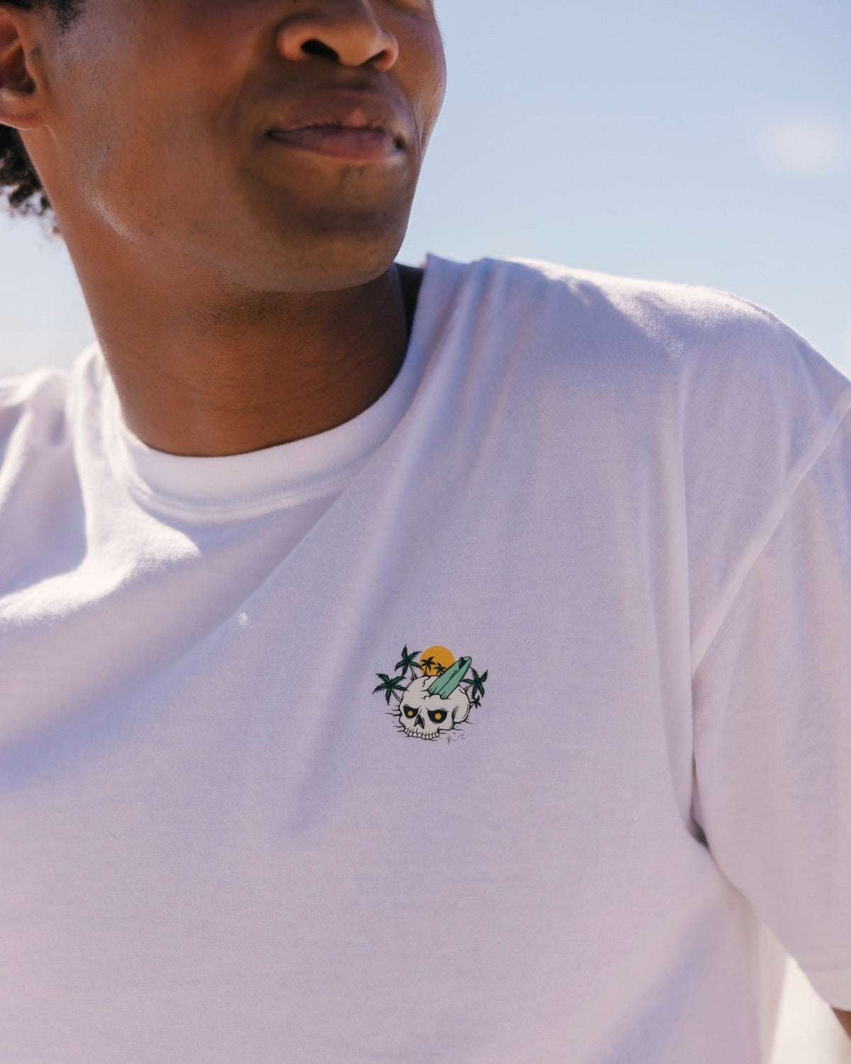 A close-up of a man wearing a white crewneck t-shirt with a small colorful embroidered crest on the left chest, set against a bright, sunlit outdoor background.
