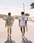 Two men walking away on a sunny beach boardwalk, each carrying a surfboard. The man on the left wears a light olive green graphic t-shirt and patterned shorts, while the man on the right wears a white graphic t-shirt and light gray shorts.