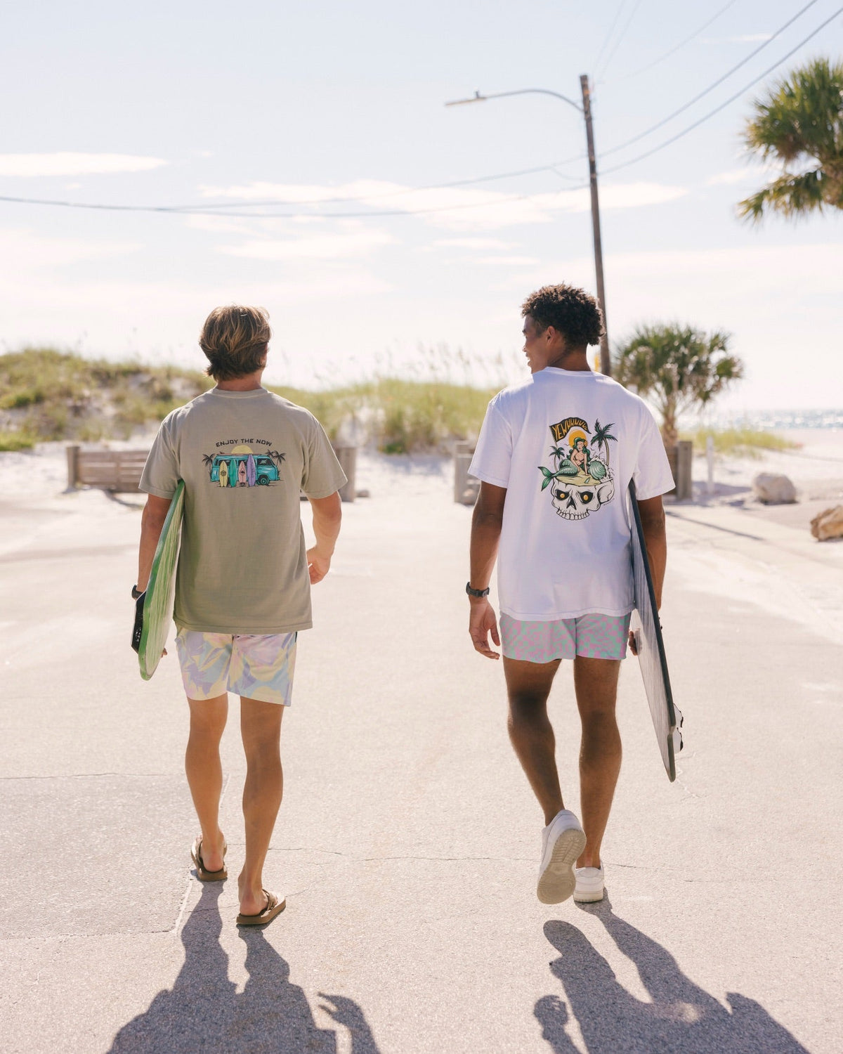 Two men walking away on a sunny beach boardwalk, each carrying a surfboard. The man on the left wears a light olive green graphic t-shirt and patterned shorts, while the man on the right wears a white graphic t-shirt and light gray shorts.