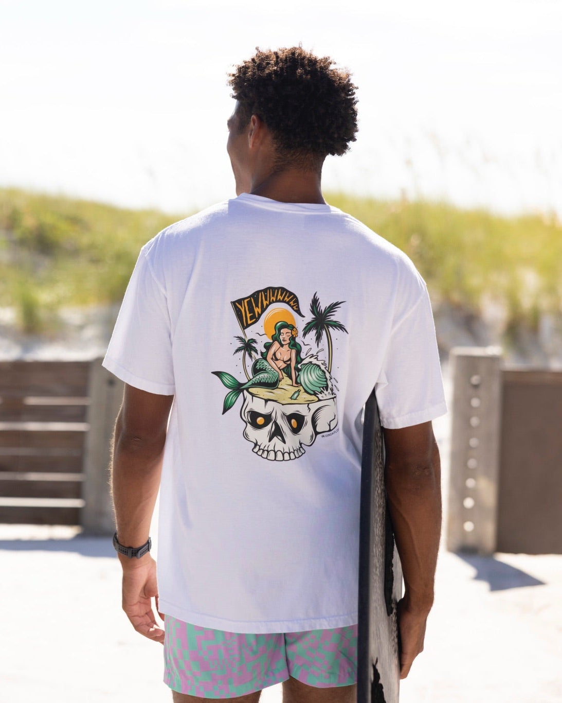 Two men seen from behind walking outdoors on a sunny beach path, each wearing a short-sleeve graphic T-shirt. The man on the left wears an olive-beige shirt with a colorful surfboard graphic and text on the back, and the man on the right wears a white shirt with a large multicolor beach-themed graphic.