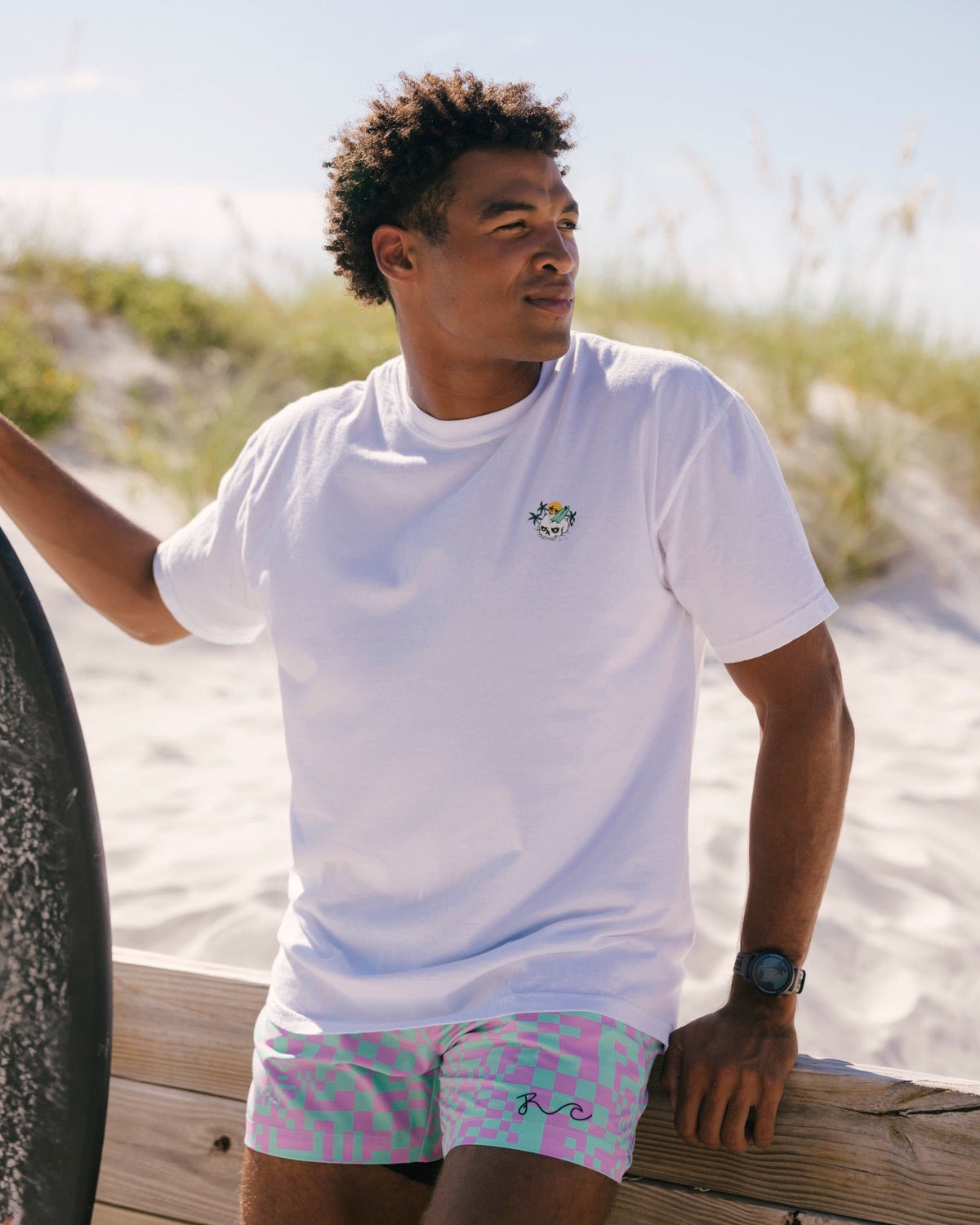A man wearing a white crewneck t-shirt and pastel patterned swim trunks, leaning on a wooden board in a sunny beach setting with sand dunes and grass in the background.
