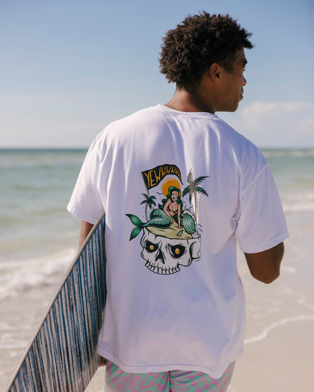 A man standing on a beach, viewed from behind, wearing a white t-shirt with a large graphic print on the back and patterned swim trunks, holding a surfboard.